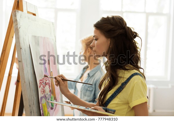 art school, creativity and\
people concept - woman with easel, palette and brush painting at\
studio