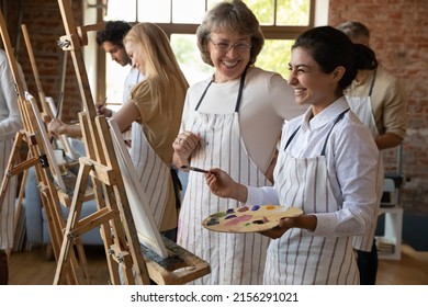 Art school student and older teacher laugh during painting lesson. Indian woman holds palette, paintbrush stand near easel enjoy hobby, artistic practice and talk with tutor. Group art-class concept - Shutterstock ID 2156291021