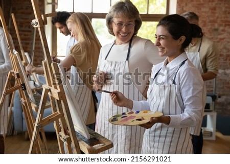 Art school student and older teacher laugh during painting lesson. Indian woman holds palette, paintbrush stand near easel enjoy hobby, artistic practice and talk with tutor. Group art-class concept