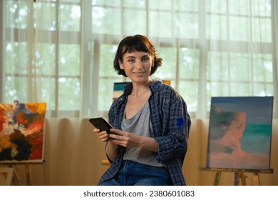 In the art studio beautiful and charismatic artist woman taking some selfies pictures with her smartphone while sitting on the chair - Powered by Shutterstock