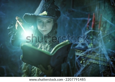 Art of witchcraft. Beautiful curly-haired sorceress in a black hat makes a spell with a magical book and a wand in a dark old castle covered in cobweb. Magic Halloween spells.