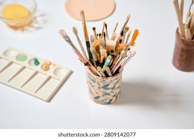 Art workshop table setup. Handmade ceramic cup holding assorted paint brushes for clay decoration. Blurry palette and sponge in the background of pottery class