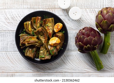 Artichoke or carciofi vegetable, raw and cooked. Artichokes boiled and roasted in olive oil with garlic, garnished with parsley and lemon. White wooden table. Top view.  
