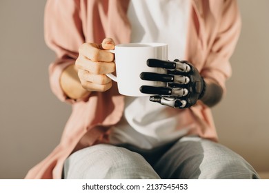 Artificial bionic female hand with mechanical metal fingers holding cup of hot drink in white cup. Woman training new skills after surgery of arm amputation. Cyborg people. Cybernetics, innovations