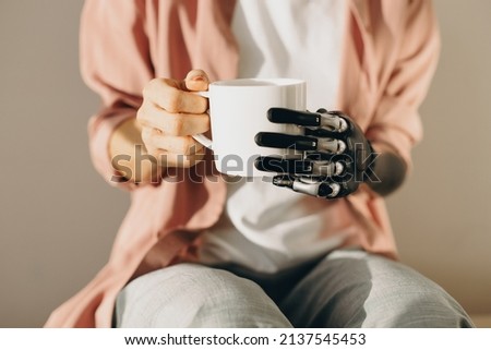 Artificial bionic female hand with mechanical metal fingers holding cup of hot drink in white cup. Woman training new skills after surgery of arm amputation. Cyborg people. Cybernetics, innovations