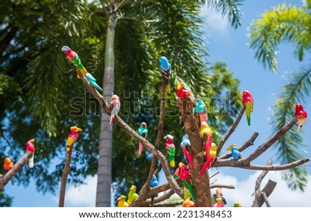 Artificial colorful parrot birds decorated on tree with blur foliage bokeh against blue sky in zoo at summer.