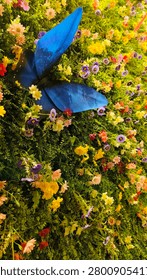 Artificial Flower Wall Backdrop with Large Blue Butterfly Decoration Colorful Floral Background. A vibrant artificial green wall backdrop decorated with numerous small, multi-colored flowers and a large, decorative blue butterfly with open wings.