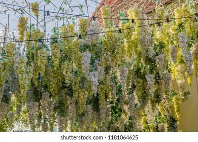 Artificial flowers hanging from string from thin rope netting with small electrical lights on a sunny day.