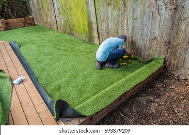 Artificial grass being installed, added next to wooden decking. It has been cut to size and rolled out and laid and is being nailed down. the installer has a hammer and nails.