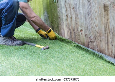 Artificial grass being installed. It has been cut to size and rolled out and laid and is being nailed down along a fence. The installer has a hammer and nails.
