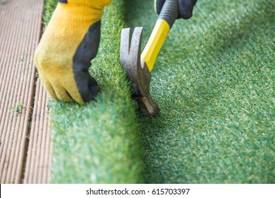 Artificial grass, turf installation alongside decking. A hammer in being used to nail the turf into place. A yellow work glove can be seen.