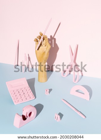 Artificial hand holding a pencil with office equipment on two tone pastel blue and pink background. Creative back to school or office abstract concept.