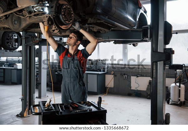 Artificial and natural lighting.\
Man at the workshop in uniform fixes broken parts of the modern\
car.