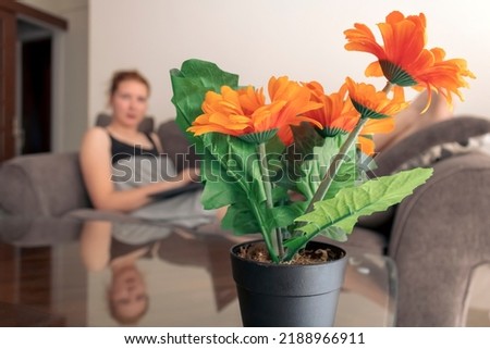 Artificial orange flowers in pot on glass coffee table in front of blurred figure of young woman lying on sofa