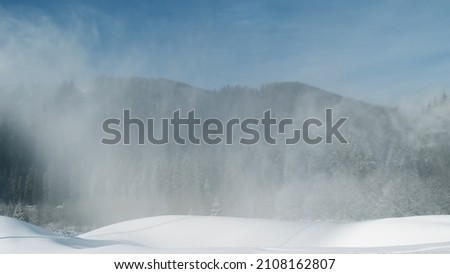 Artificial snow falls on background of spruce forest in mountains at ski resort. White snow-covered mountain slope for skiing on sunny frosty day in winter. Working snow cannon, snowgun on blue sky
