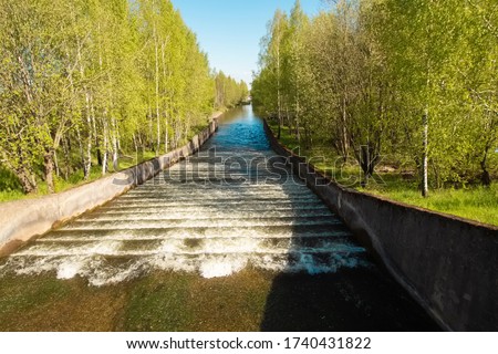 Artificial waterfall cascade under water stairs on a river channel in Zaslavl Belarus. Bright sunny weather, green grass and blue sky and fast water stream.