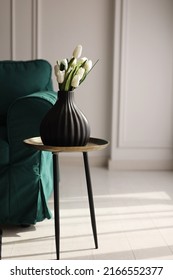 artificial white tulips and green leaves in a black vase on the table in home interior