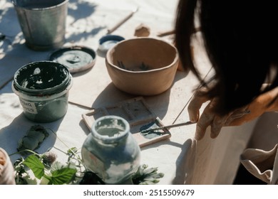 An artisan applies green paint to a canvas using a brush, surrounded by various natural materials and tools on a sunlit table in a garden setting. - Powered by Shutterstock