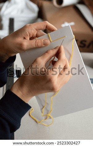 Artisan binding a book using traditional Japanese bookbinding techniques with yellow thread and white paper. The intricate stitching process, minimalist design, handmade craftsmanship.