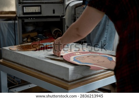 Artisan placing handcrafted hot enamel metal plates depicting orthodox icons of Virgin Mary and Jesus Christ onto marble table in craft workshop.