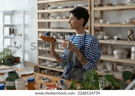 Artisan tattooed girl drink coffee and looking at tasty bun lustily in art studio during break from producing handmade pottery for handicraft shop. Potter woman has lunch with tea on workplace