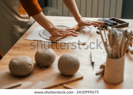 Artisan woman in lighted pottery studio, rolling out clay for modelling. Pottery studio with a variety of ceramics and a focused artisan