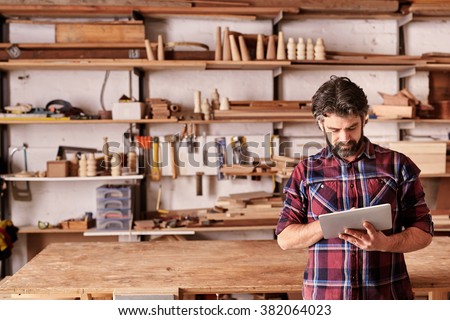 Artisan woodwork studio with shelving holding pieces of wood, with a carpenter standing in his workshop using a digital tablet