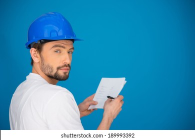 Artisan Or Workman Taking Notes Standing In Front Of A Blue Wall With A Pen And Paper In His Hands Turning To Look At The Camera With A Serious Thoughtful Expression