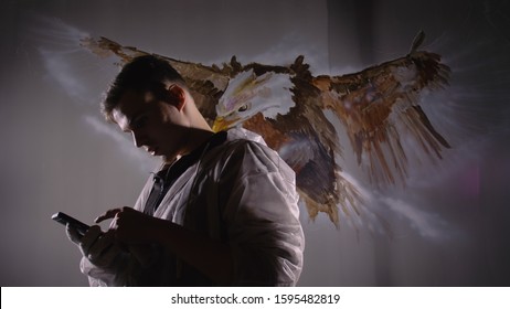 Artist designer draws an eagle on wall. Craftsman decorator paints picture with acrylic oil color looking at sketch in phone. Painter painter dressed in paint coat. Indoor. Dark magic cinematic look. - Shutterstock ID 1595482819