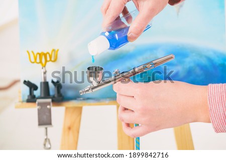 Artist fills the airbrush canister with blue paint. An easel with a painting is visible in the background