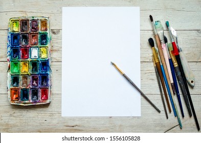 Artist paint brushes and watercolor paintbox on wooden background with empty sheet of paper. Instruments and tools for creative leisure. Paintings art concept mockup. Top view. Copy space. Flat lay.