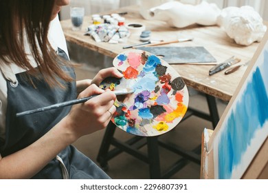 The artist with a palette and a brush in her hands. Close-up. White T-shirt and apron. Portrait. Creative person concept. Art materials in the background. Workshop interior.  - Powered by Shutterstock