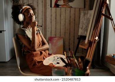 Artist sitting in cozy studio wearing headphones, holding paintbrush under chin while deep in thought, surrounded by art supplies and canvases - Powered by Shutterstock