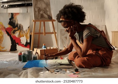 Artist sitting on floor painting with various art supplies around. Abstract and portrait paintings on easel and wall in background enhancing creative atmosphere - Powered by Shutterstock