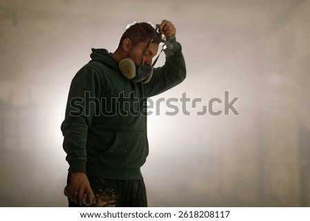 An artist stands in a dimly lit studio, wearing a gas mask while holding spray paint. He appears focused and contemplative, preparing for a painting session that emphasizes creativity.