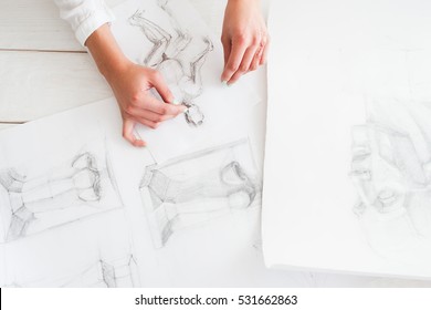 Artist workplace with pencil pictures flat lay. Top view on table with different sketches and female artist hands, drawing female silhouette. Art, craft, creativity, inspiration concept