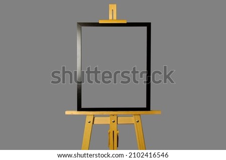 An artistic easel with a picture in black frame isolated on a gray background. The information frame is mounted on a tripod.