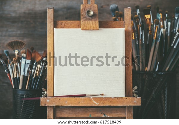 Artistic equipment\
in a artist studio: empty artist canvas on wooden easel and paint\
brushes Retro toned\
photo.