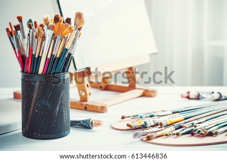 Artistic equipment: easel, paint brushes, tubes of paint, palette and paintings on work table in a artist studio.