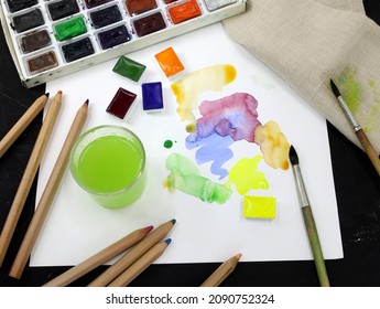 Artistic paints, a glass of water, pencils and a sheet of paper. Watercolor paints in a plastic container and colored paint strokes on a sheet. Brush strokes of paint on watercolor paper.