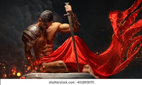 Artistic portrait of muscular male in red waving fabric with fire sparks holding sword.