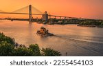 Artistic view of evening in Savannah, Georgia, as tourists gather on the river bank and a tug boat makes a wake heading into the golden sky under the Talmadge Memorial Bridge.