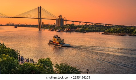 Artistic view of evening in Savannah, Georgia, as tourists gather on the river bank and a tug boat makes a wake heading into the golden sky under the Talmadge Memorial Bridge.