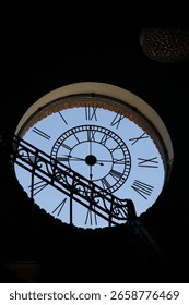 Artistic view of a large round clock with Roman numerals, seen from the interior through a silhouetted staircase. 