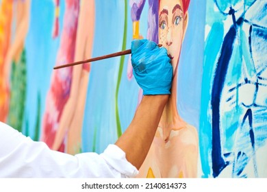 Artist's Hand With Paintbrush Painting Colorful Picture On Canvas At Outdoor Art Festival. Woman Painter In Blue Gloves Draws Surreal Picture With Paintbrush, Beautiful Art Performance