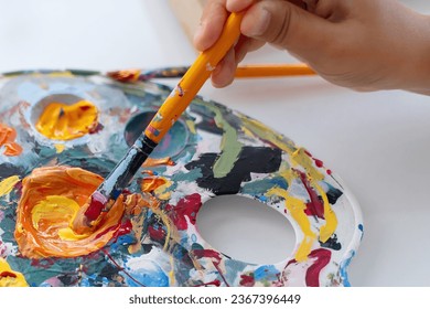 Artist's hand picks up acrylic or oil paint from palette on brush. Closeup paintbrush picking orange color from an artist palette. Colorful image from an artist’s studio or a school showing creative  - Powered by Shutterstock