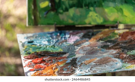 An artist's wooden palette with vibrant oil paint colors sits in focus against a blurred natural landscape, capturing the essence of painting en plein air. - Powered by Shutterstock