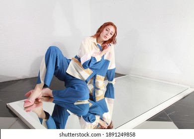 Artsy Fashion Image Of Ginger Woman With Pale Skin Wearing Summer Blue And White Pants Suit And Lying On A Mirror In A Spacious Room. Studio Catalog Shooting. Future Of Fashion. Trendy Summer Clothes.