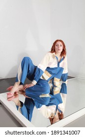 Artsy Fashion Image Of Ginger Woman With Pale Skin Wearing Summer Blue And White Pants Suit And Lying On A Mirror In A Spacious Room. Studio Catalog Shooting. Future Of Fashion. Trendy Summer Clothes.