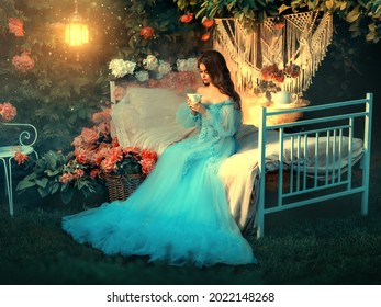 Artwork Fairy Tale Princess Sits On Vintage Bed. Fantasy Woman Holding Cup In Hands Drinking Tea. Long Wave Hair. Blue Vintage Luxury Dress. Backdrop Blooming Flowers, Old Lamp. Fantasy Woman Queen